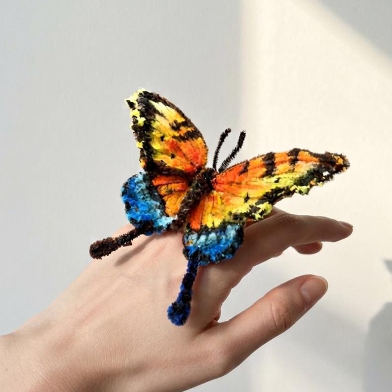 Puede incluir: Un anillo con forma de mariposa hecho de limpiapipas difusos. Las alas presentan un degradado vibrante de amarillo, naranja y negro, con acentos azul brillante en las secciones inferiores. Dos antenas finas y oscuras sobresalen del cuerpo.