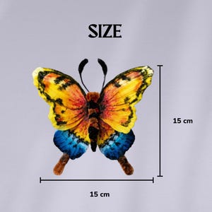Puede incluir: Un adorno de mariposa con alas amarillas, naranjas y azules con marcas negras y dos antenas. El texto SIZE est&aacute; centrado sobre la mariposa. Las l&iacute;neas de medici&oacute;n indican una altura de 15 cm y una anchura de 15 cm para la mariposa.