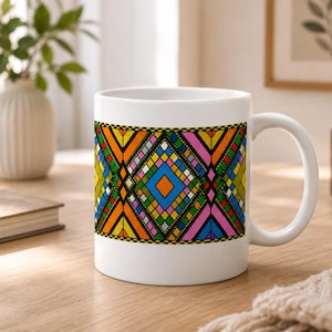 Könnte beinhalten: Ein weißer Keramikbecher mit einem horizontalen Band aus bunten geometrischen Mustern. Das Design besteht aus sich wiederholenden Rauten und Dreiecken in leuchtenden Orange-, Blau-, Rosa-, Grün- und Gelbtönen, die schwarz auf dem Weiß umrandet sind.