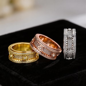 May include: Three ornate rings in gold, rose gold, and silver. Each ring features a gear-like design with embedded gemstones. The rings are displayed on a black surface, showcasing their intricate details and varying metallic finishes.