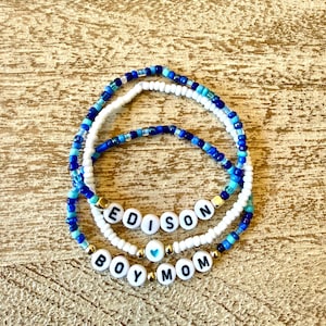 May include: Three beaded bracelets in shades of blue and white. One bracelet spells "EDISON" in white letter beads, and another spells "BOY MOM". The bracelets are on a light brown wooden surface.