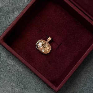 Può includere: Un pendente in oro con una grande pietra preziosa di colore chiaro, taglio quadrato, è esposto all'interno di un portagioie bordeaux. La scatola è aperta, rivelando il pendente contro un interno in velluto rosso scuro. Il pendente è su uno sfondo scuro.