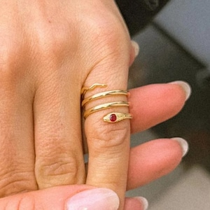 May include: A gold ring in a spiral design, encircling a finger. The ring has a small red gemstone. The jewellery item is a ring.