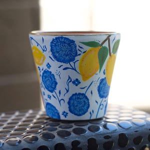 May include: A white ceramic plant pot featuring a hand-painted pattern of bright yellow lemons with green leaves and vibrant blue floral blossoms. The pot has a tapered shape with a smooth finish and a visible terracotta rim at the top edge.