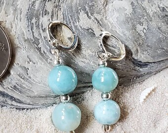 May include: A pair of dangle earrings featuring two light blue, round beads connected by silver-colored metal. The earrings have a lever-back closure. The earrings are displayed on a sandy surface with a seashell.