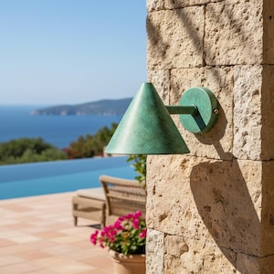 May include: A green outdoor wall light with a conical shade. The fixture is mounted on a stone wall, with a view of the sea and sky in the background. The light has a circular base and a straight arm. The light is made of metal.