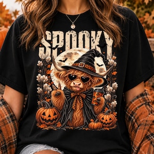 May include: A black t-shirt featuring a graphic of a fluffy highland cow wearing a witch hat, heart-shaped sunglasses, and a cape. The cow holds a coffee cup and makes a peace sign. The text SPOOKY appears above, with pumpkins and a moon.