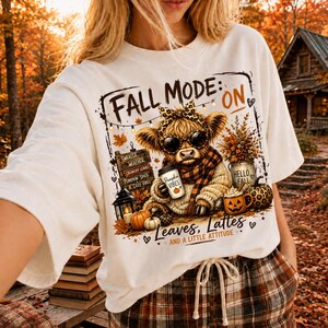 May include: A white t-shirt featuring a graphic of a Highland cow wearing sunglasses and a leopard print bow. The design includes text reading FALL MODE: ON, PUMPKIN VIBES, HELLO FALL, and Leaves, Lattes AND A LITTLE ATTITUDE, with autumn motifs.