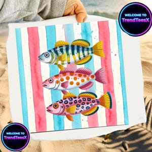 May include: A white beach towel with three vibrant fish designs set against a backdrop of pink and blue watercolour stripes. The fish are intricately patterned with yellow fins. The towel has the text "WELCOME TO TrendTeesX" in a circular design.