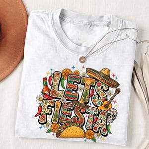 May include: White t-shirt with the words "Let's Fiesta" in a colorful floral design. The design includes a sombrero, guitar, taco, hot sauce, and a sugar skull. A gold necklace is draped over the shirt.