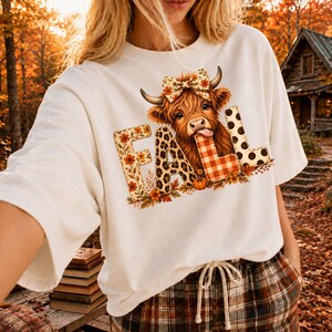 May include: A white t-shirt featuring a graphic of a Highland cow with a floral bow on its head. The cow is positioned behind the word FALL, where each letter has a different pattern: floral, leopard print, orange plaid, and black polka dots.