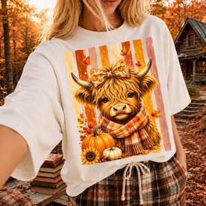 May include: A white t-shirt featuring a graphic of a fluffy Highland cow wearing a plaid scarf and a matching bow. The cow is positioned above a large orange pumpkin and a smaller white pumpkin, set against a background of vertical autumn stripes.