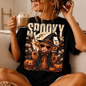 May include: A black t-shirt featuring a graphic of a fluffy highland cow wearing a witch hat and heart-shaped sunglasses. The cow holds a coffee cup and makes a peace sign. The design includes pumpkins, bats, and the text SPOOKY in large letters.