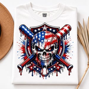 May include: A white t-shirt with a Comfort Colors label featuring a graphic of a skull painted like an American flag, positioned in front of two crossed baseball bats. The design includes red, white, and blue paint drips, stars, and a baseball.