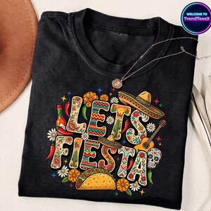 May include: Black t-shirt with a colorful "Let's Fiesta" graphic. The design includes sombreros, a guitar, a taco, flowers, and a sugar skull. A silver necklace with a pendant is draped across the shirt.
