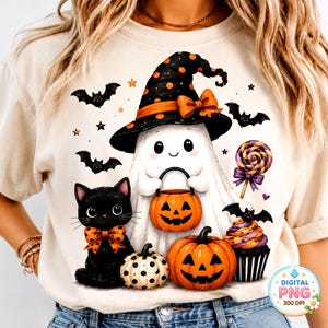 May include: A cream-colored t-shirt featuring a graphic of a white ghost wearing a black witch hat with orange polka dots and a bow, holding an orange jack-o'-lantern bucket. Beside the ghost is a black cat with an orange bow, pumpkins, bats, a lollipop, and a cupcake.