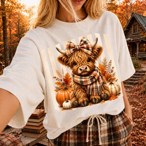 May include: A white t-shirt featuring a graphic of a fluffy brown highland cow wearing a plaid bow and scarf, surrounded by orange pumpkins and autumn leaves. The design is set against a background of vertical light-colored stripes.