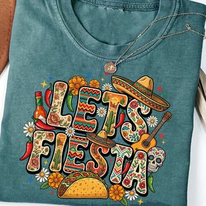 May include: Teal-colored t-shirt with a colorful "Let's Fiesta" graphic. The design includes a sombrero, guitar, taco, chili peppers, flowers, and a sugar skull. A gold necklace with a pendant is also visible.