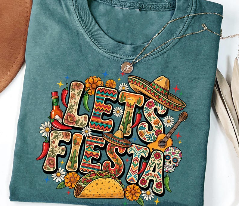 May include: Teal-colored t-shirt with a colorful "Let's Fiesta" graphic. The design includes a sombrero, guitar, taco, chili peppers, flowers, and a sugar skull. A gold necklace with a pendant is also visible.