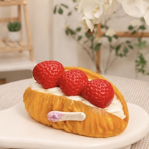 May include: A golden-brown, textured pastry shell filled with white cream and topped with three bright red strawberries. A small, light-colored oval tag is attached to the side of the pastry, featuring a small purple graphic and the text "icecream".