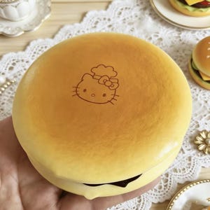 May include: A round, golden-brown pancake-shaped object featuring a brown line-art illustration of the Hello Kitty character wearing a chef hat. The surface has a smooth, matte texture with a dark brown filling visible along the bottom edge.