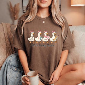 May include: A brown t-shirt with a playful illustration of four geese, each wearing a pink bow and carrying accessories. The geese are shown with glasses, a book, a coffee cup, and a pencil. The text "Off To Actively Monitor" is printed below.