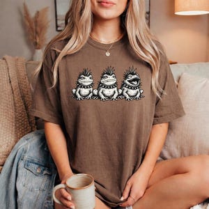 May include: Brown t-shirt featuring a graphic of three cartoon frogs with spiky mohawks. The frogs are in a row, with one frog's mouth open. The t-shirt is a solid brown color.