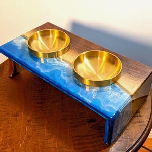 May include: A rectangular elevated pet feeder stand featuring a dark brown wood surface combined with a vibrant blue and white resin wave design. The stand includes two circular, polished gold-toned metal bowls set into the top surface.