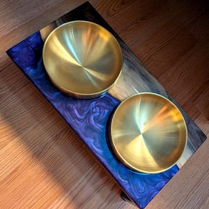 Peut inclure: Un support rectangulaire pour animaux avec deux bols circulaires en métal doré insérés dans une base en bois naturel et une incrustation en résine marbrée aux reflets violets et bleus. Les bols sont lisses et réfléchissants.