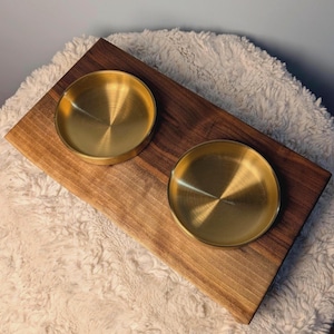 May include: A rectangular dark brown wooden board featuring two circular, polished gold-toned metal bowls embedded into the surface. The wood displays a natural grain pattern, and the metal bowls have a smooth, reflective finish with a circular texture.