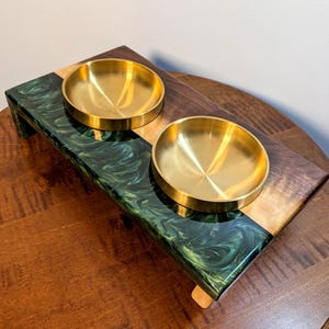 Peut inclure: Un support surélevé rectangulaire pour animaux, composé de bois brun foncé et d'une section en résine aux motifs tourbillonnants vert foncé et noir. Deux bols circulaires en métal doré poli sont insérés sur le dessus du support.