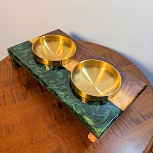 May include: A rectangular elevated pet feeder stand featuring a combination of dark brown wood and a swirling dark green resin section. Two circular, polished gold-tone metal bowls are set into the top surface of the stand, which is supported by four short legs.