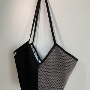 Könnte beinhalten: Eine Tragetasche mit geteiltem Design, bei der eine Seite aus einfarbigem schwarzem Stoff und die andere aus weißem Netzmaterial mit sechseckigem Muster besteht. Die Tasche hat zwei schwarze Schulterriemen und eine V-förmige Öffnung.