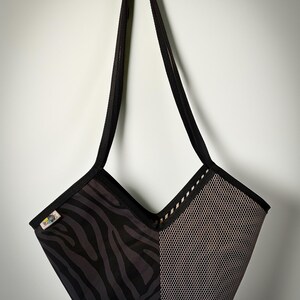 Könnte beinhalten: Eine Tragetasche mit geteiltem Design, bestehend aus einem schwarz-dunkelgrauen Zebramuster auf der einen Seite und einem strukturierten hellgrauen Netzstoff auf der anderen. Die Tasche hat eine V-förmige Öffnung, schwarze Ränder und zwei lange Tragegurte.