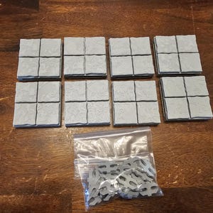 May include: Eight grey, square, textured tiles arranged in two rows, each tile divided into four smaller squares. A clear plastic bag contains small, silver-coloured connector pieces. The tiles appear to be for a tabletop game.