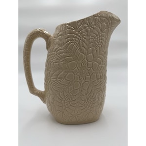 May include: A beige ceramic pitcher featuring a raised, intricate lace-like pattern covering the entire exterior surface. The pitcher has a curved handle on the left side and a wide, pointed pouring spout at the top rim, with a glossy finish.
