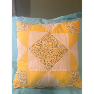 May include: A square throw pillow featuring a patchwork design with a central diamond and corner accents in a yellow and white floral print. The remaining sections consist of solid yellow and white fabric panels arranged in a geometric star pattern.