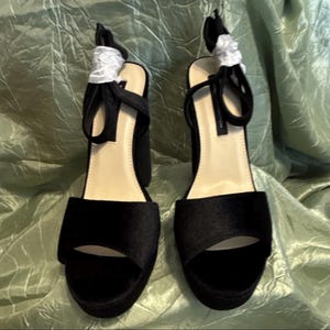 May include: Pair of black platform sandals with open toes and ankle straps. The sandals have thick block heels and are wrapped in a soft, textured fabric. The straps are tied with white ribbon.