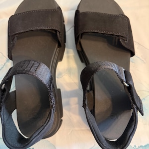 May include: A pair of black sandals with adjustable hook-and-loop straps. The sandals have a thick sole and a strap that goes around the ankle. The sandals are made of a soft, suede-like material.