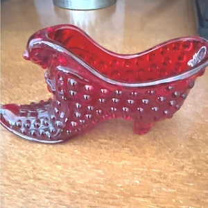 May include: A translucent ruby red glass shoe figurine featuring a textured hobnail pattern across its exterior surface. The decorative object is shaped like a high-heeled boot with an open top, showcasing a glossy finish and a distinct raised dot design.