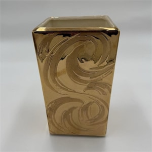 May include: A gold-colored, square vase with a glossy finish. The vase features a swirling, embossed design on its exterior. The rim of the vase is also gold. The interior of the vase is a cream color.