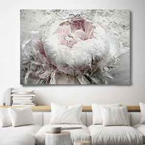 May include: A large canvas print featuring a close-up of a peony flower in shades of white and pale pink. The artwork has a textured, distressed appearance. The print hangs above a white sofa with matching pillows and two small, cylindrical side tables.