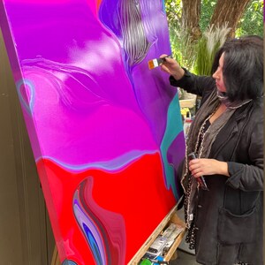 May include: An artist painting a large abstract canvas. The artwork features vibrant colors, including red, purple, and teal, with a fluid, marbled effect. The artist is using a brush to add details to the painting.