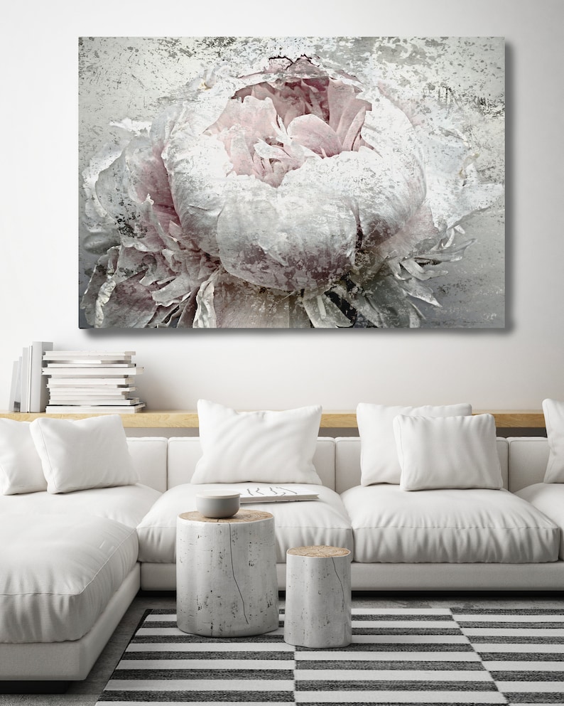 May include: A large canvas print featuring a close-up of a peony flower in shades of white and pale pink. The artwork has a textured, distressed appearance. The print hangs above a white sofa with matching pillows and two small, cylindrical side tables.