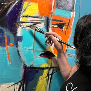 May include: A person paints a colorful abstract portrait on a canvas. The painting features bold strokes of blue, orange, yellow, and black paint. The artist is using a brush to add details to the painting.