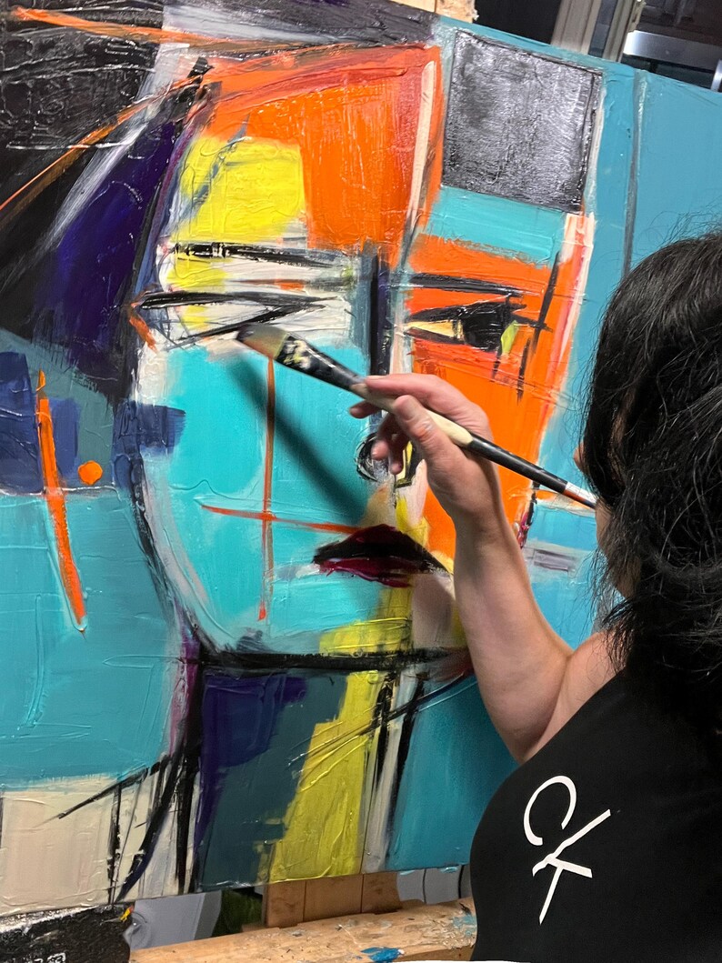 May include: A person paints a colorful abstract portrait on a canvas. The painting features bold strokes of blue, orange, yellow, and black paint. The artist is using a brush to add details to the painting.