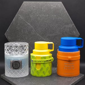May include: Three fragrance bottles: a frosted glass bottle with a diamond-patterned cap and silver label, a yellow bottle with a green leaf-patterned sleeve labeled ODYSSEY LIMONI, and an orange leather-cased bottle with a blue cap labeled ODYSSEY ELIXIR.