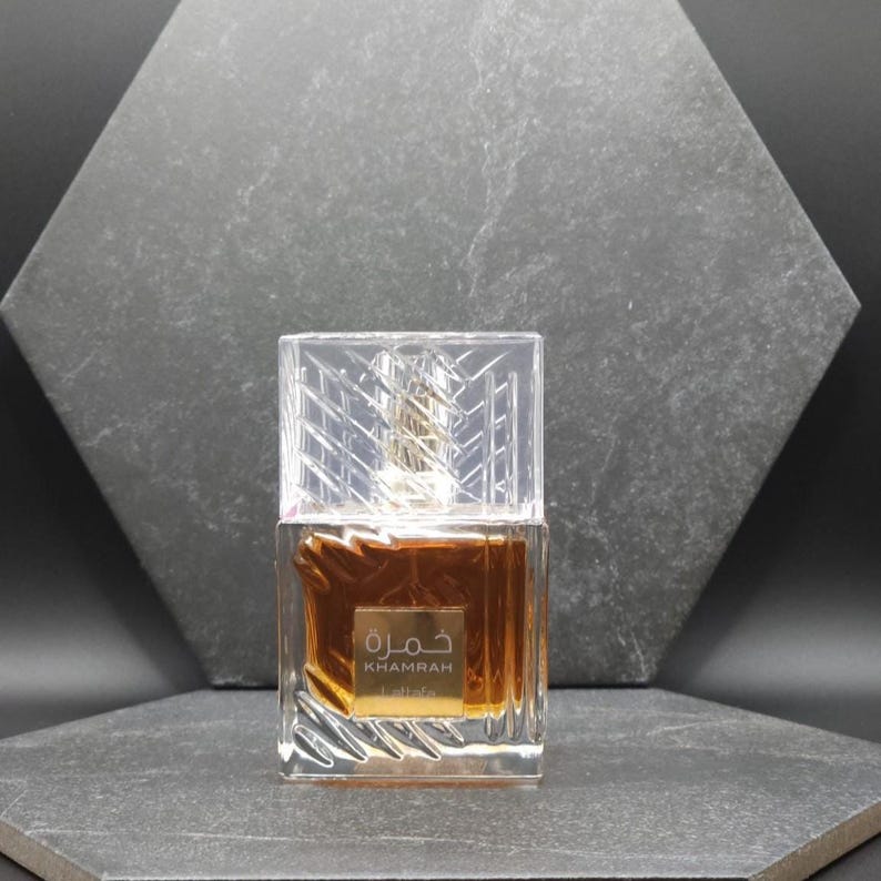 May include: A clear, rectangular perfume bottle with a gold-colored liquid inside. The bottle has a clear, geometric cap. The label reads "Khamrah" in Arabic and English. The background is a dark gray, textured surface.