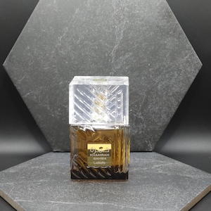 May include: A brown perfume bottle with a clear, geometric cap. The bottle has a gold label with Arabic script and the words "Khamrah Qahwa Lattafa". The background is a dark gray, textured surface.