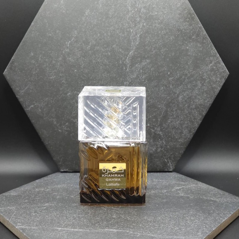 May include: A brown perfume bottle with a clear, geometric cap. The bottle has a gold label with Arabic script and the words "Khamrah Qahwa Lattafa". The background is a dark gray, textured surface.